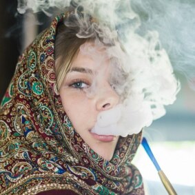 زنی با شال اروپای شرقی در حال کشیدن قلیان و بیرون دادن دود، در فضایی نیمه‌روشن و هنری